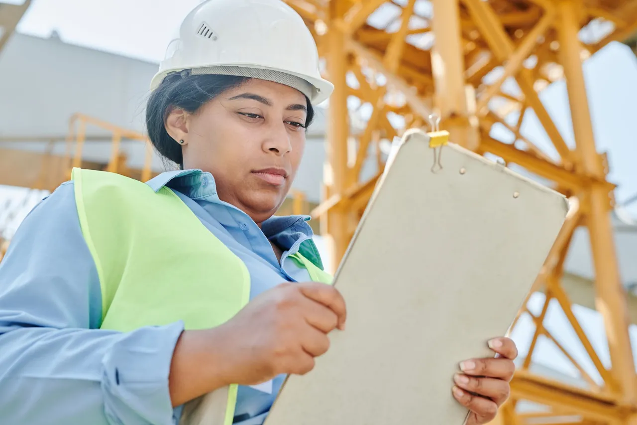 An inspector in a hard hat and safety vest reviewing a clipboard at a construction site